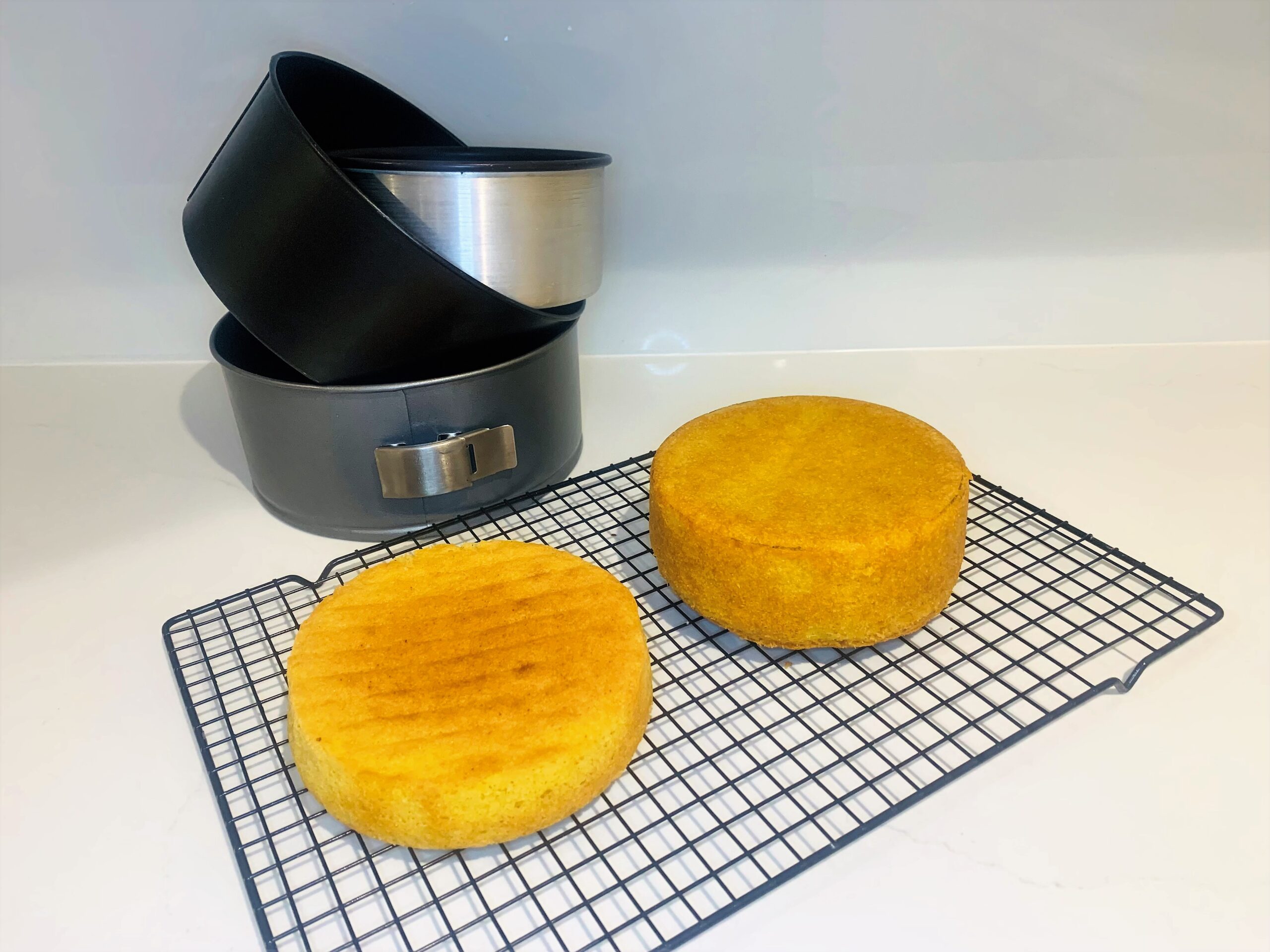 Three round baking tins and two vanilla sponge cakes cooling on a black wire cool rack.