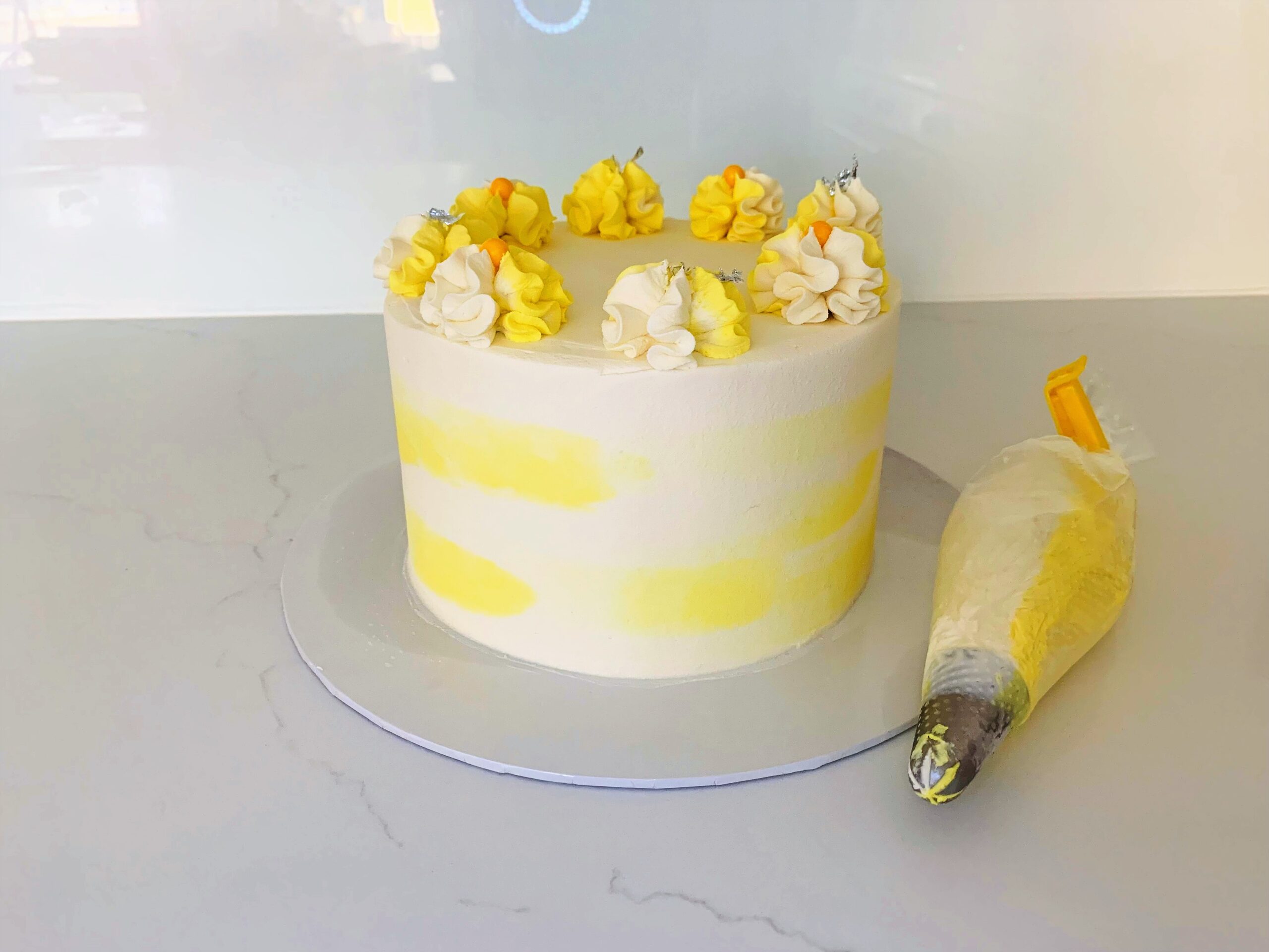 Round buttercream iced cake in yellow and white with a piping bag beside it.