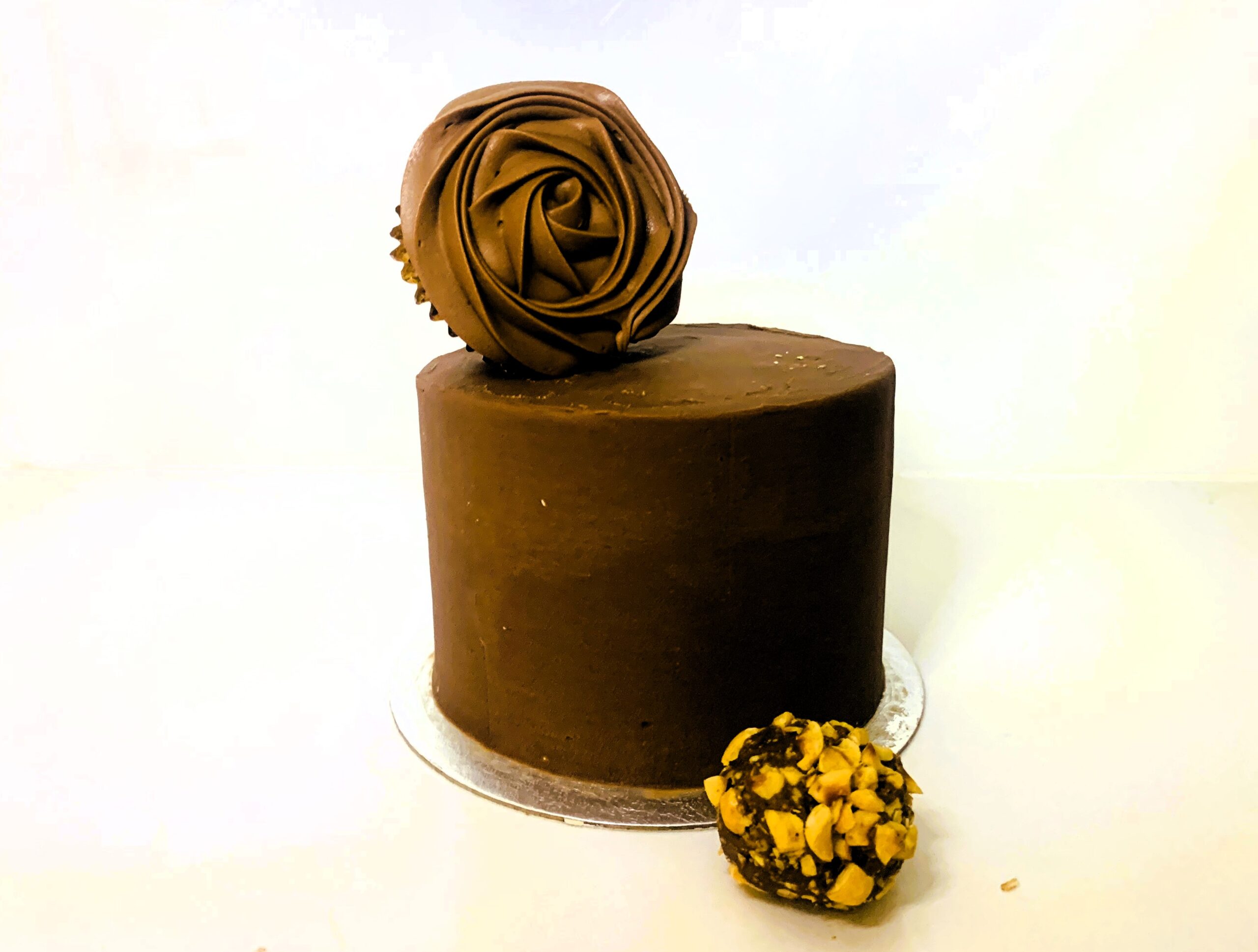 Cake covered in milk chocolate ganache with a cupcake on top that has a piped rose design in ganache and nut covered truffel chocolate