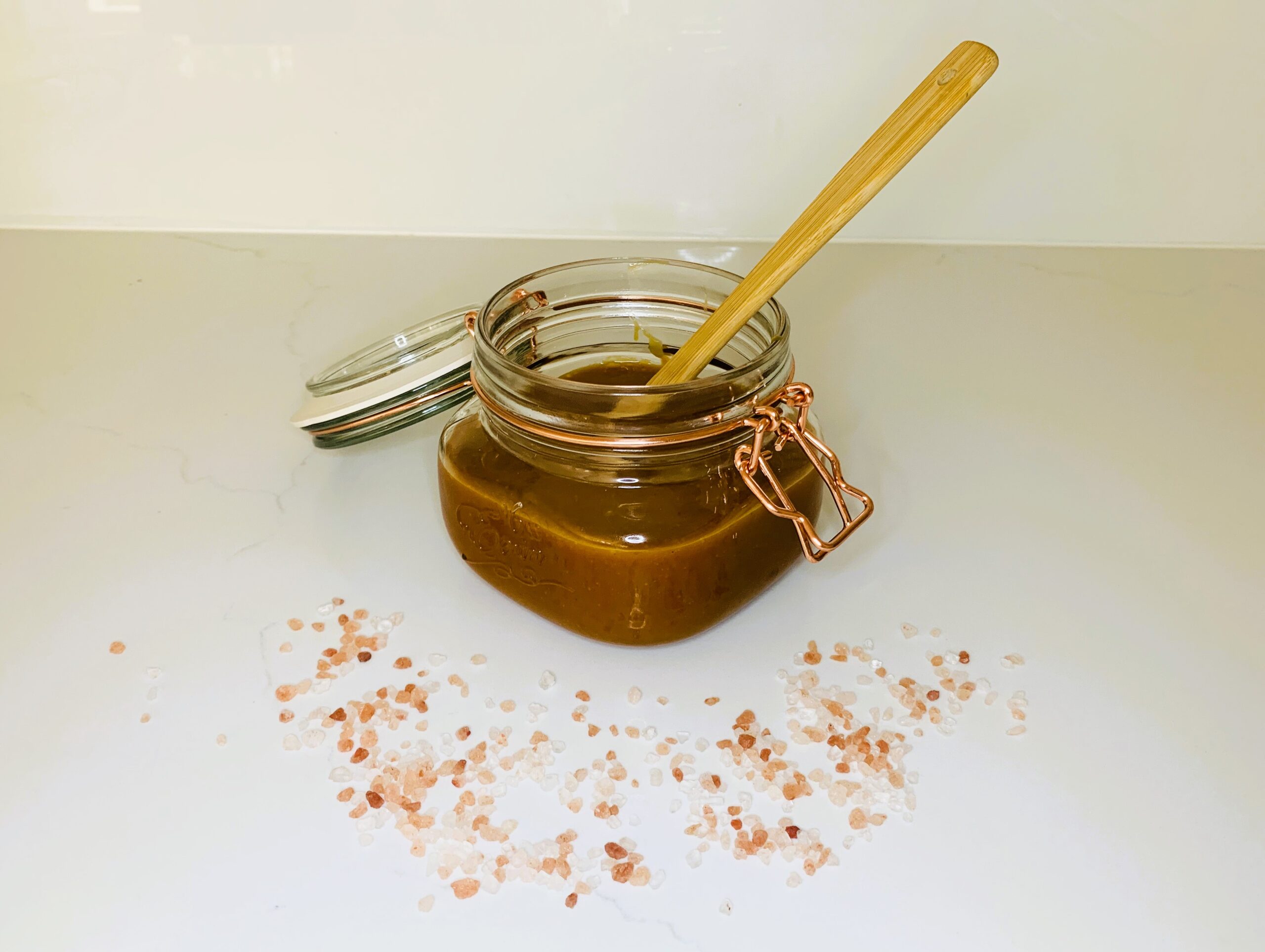 Hoemade salted caramel in a jar with a small spatula in it and surrounded by pink salt