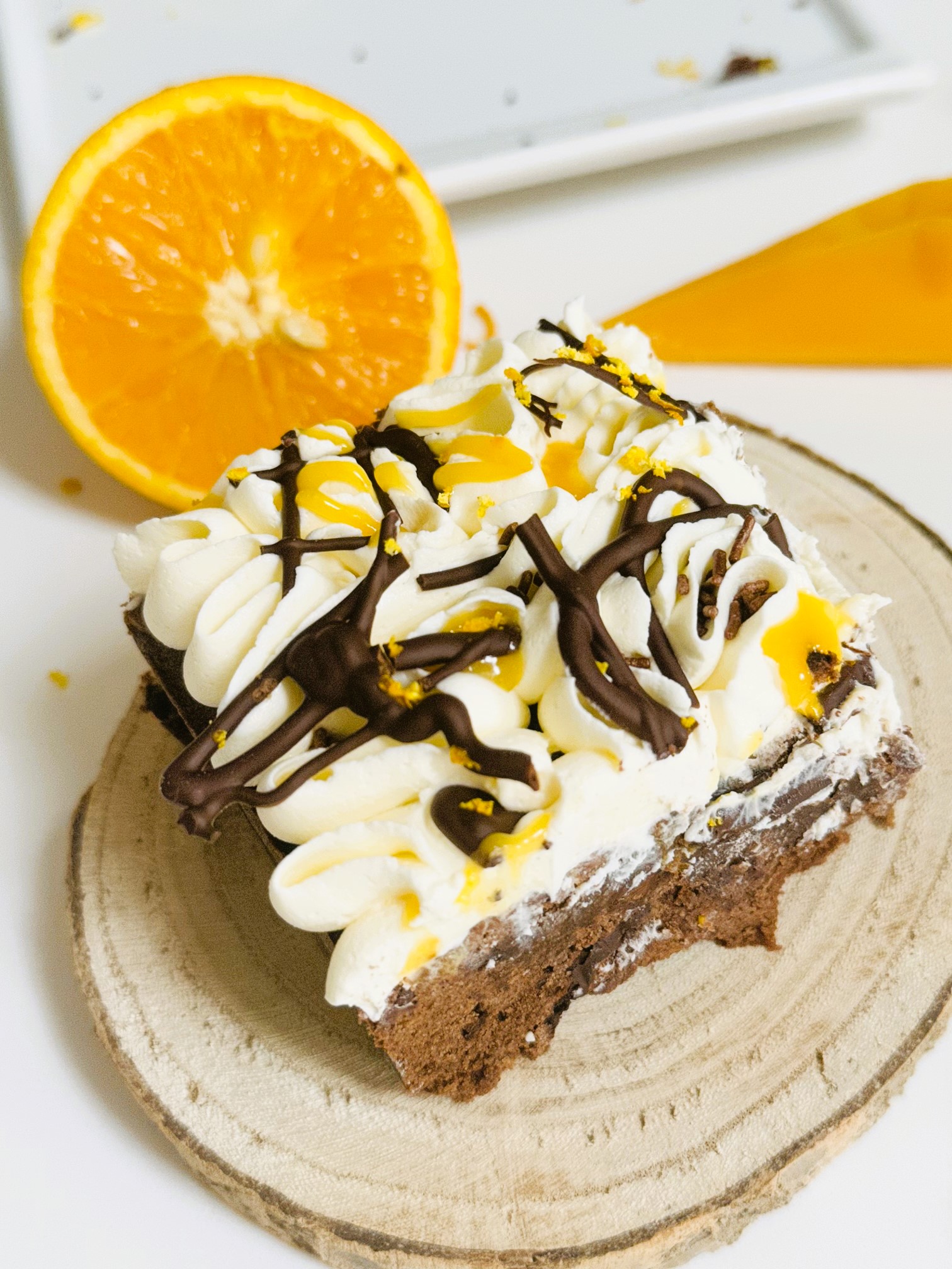 Slice of loaded dark chocolate and orange brownie that is topped with orange curd. Orange curd in a piping bag and half of a fresh orange.