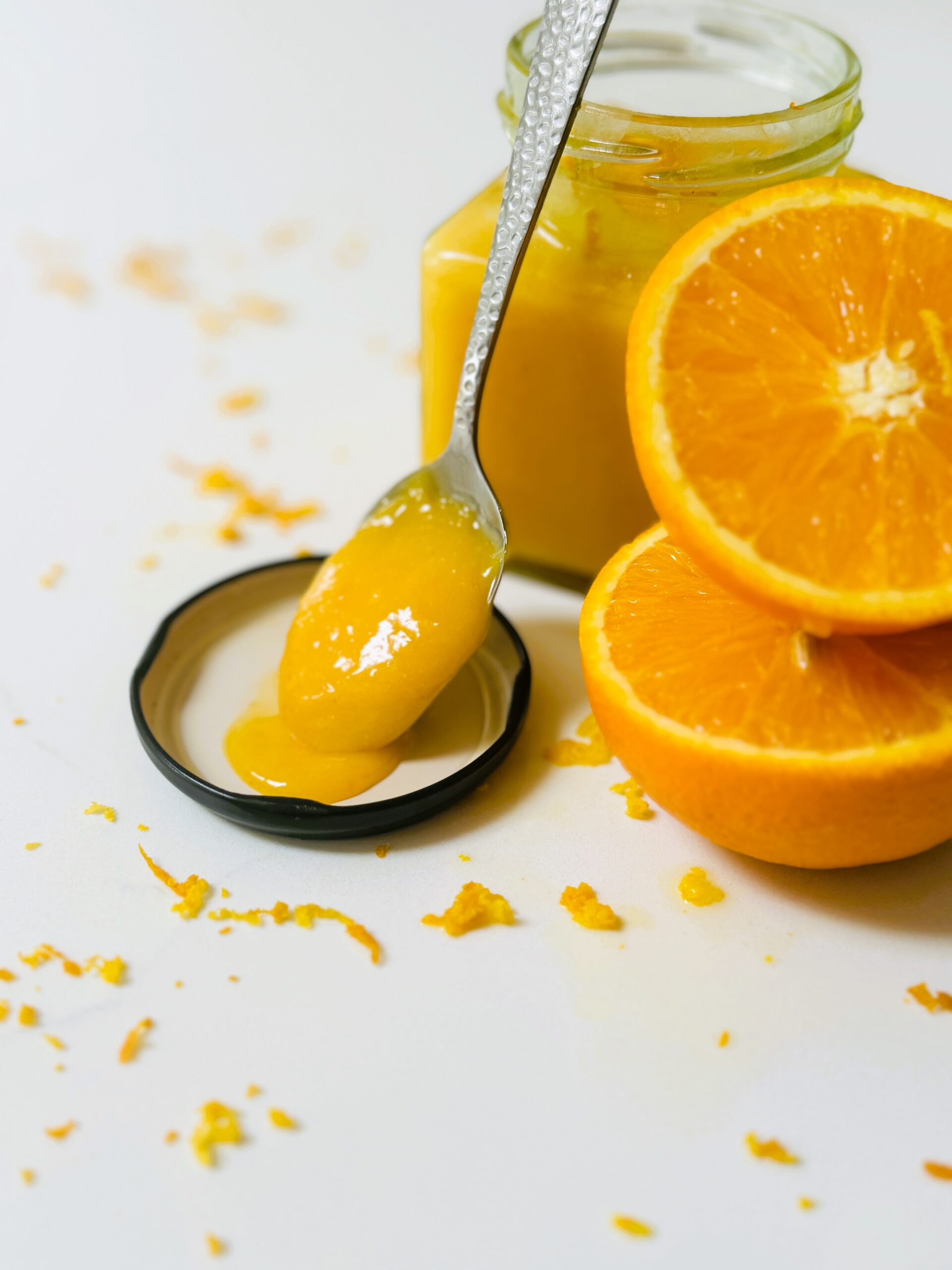 Homemade Orange Curd in a glass jar with an orange cut in half and some zest. Orange curd on a spoon slightly runnding off into jar lid.